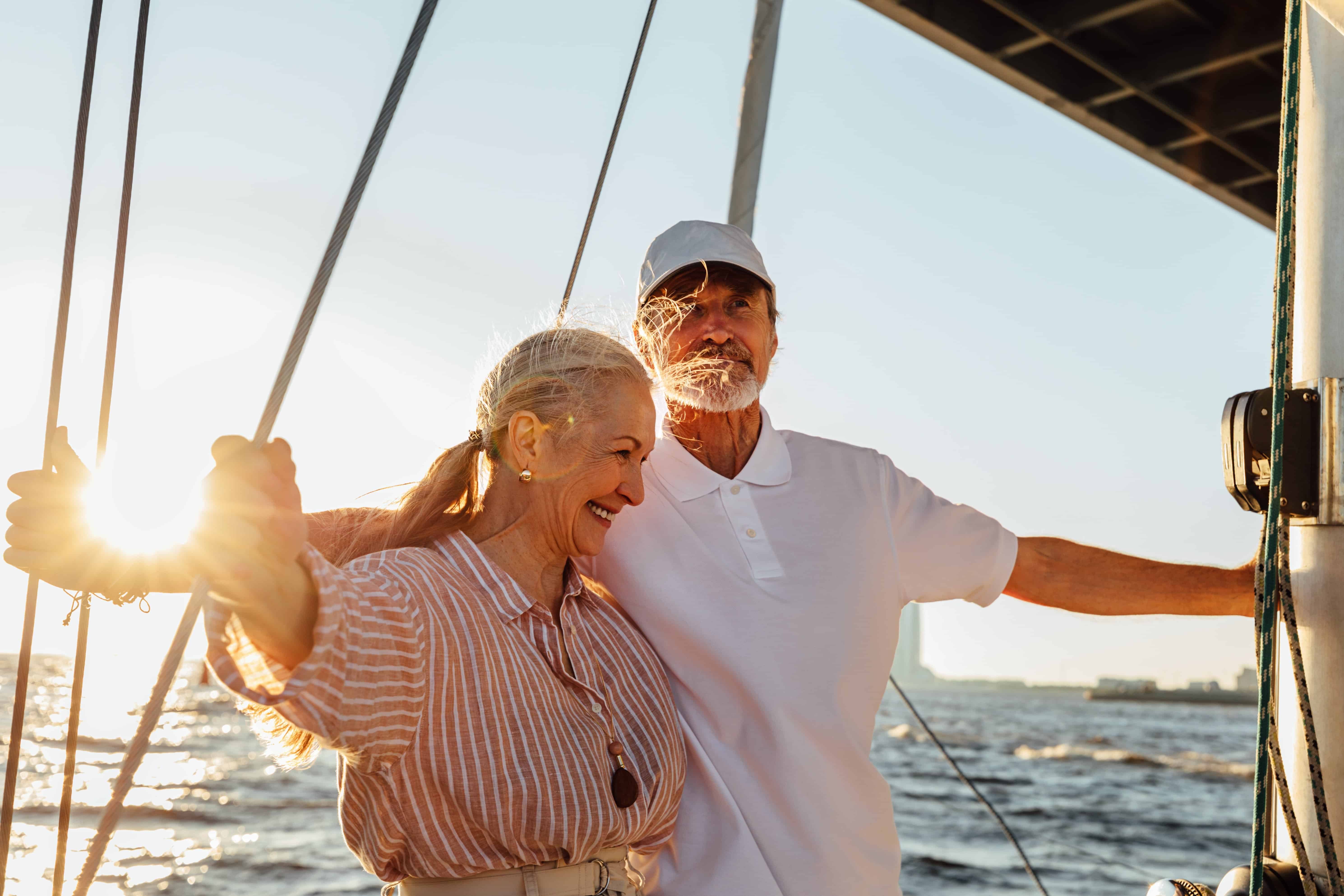 Happy senior couple enjoying a sailing trip at sunset. The woman smiles while holding onto ropes, and the man in a white polo and cap stands beside her, both relaxed and joyful on the water.