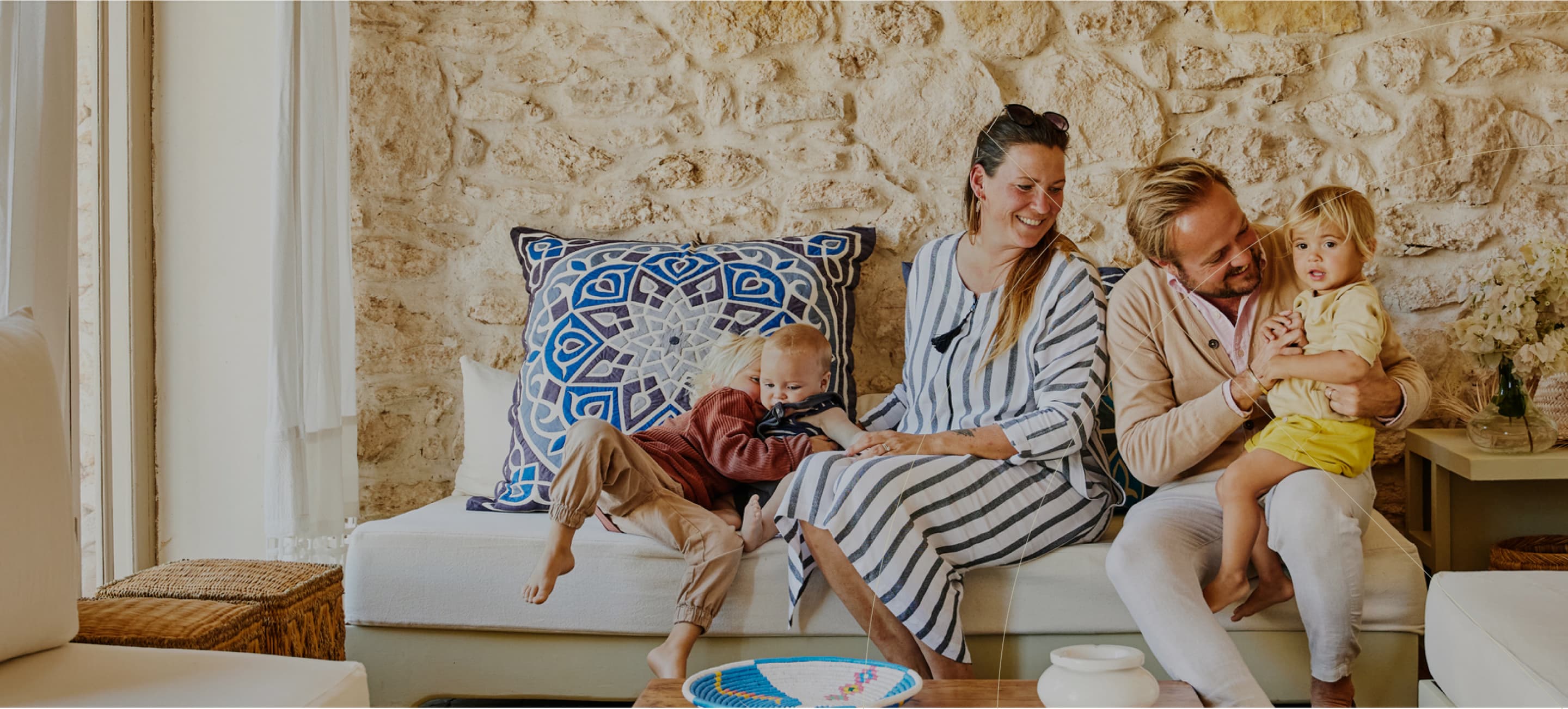 Smiling couple sitting on a sofa with their young child, in a cozy, sunlit room.