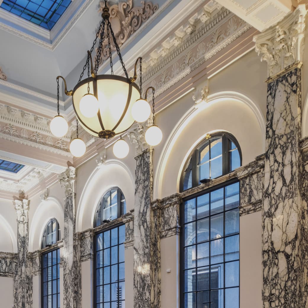 Interior of a grand building with tall arched windows, ornate marble columns, decorative molding, and a large chandelier with round lights hanging from the ceiling.