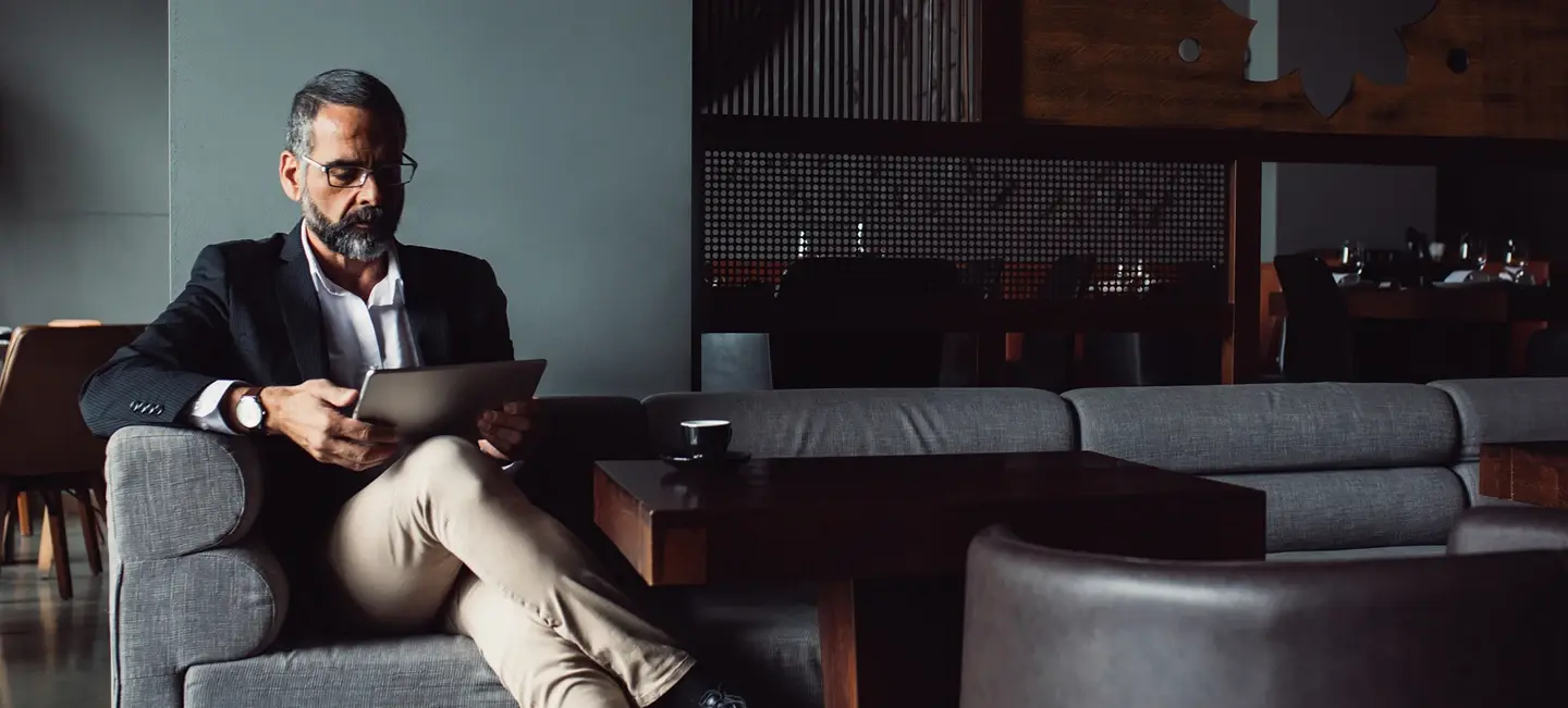 Man sitting on a sofa in a lounge, wearing a dark jacket and light pants, reading a document.