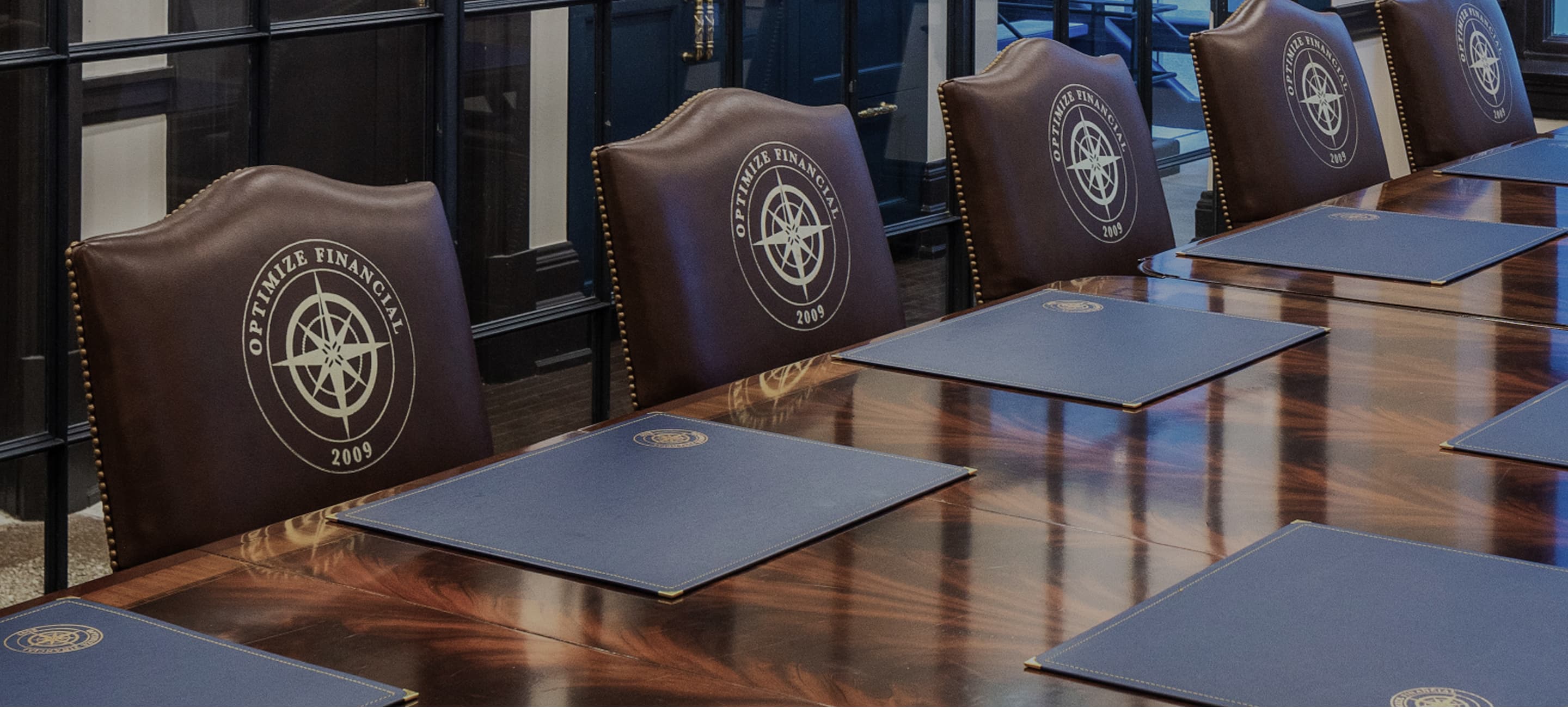 Row of brown leather chairs with logos on the backrests, arranged behind tables in a formal meeting room.