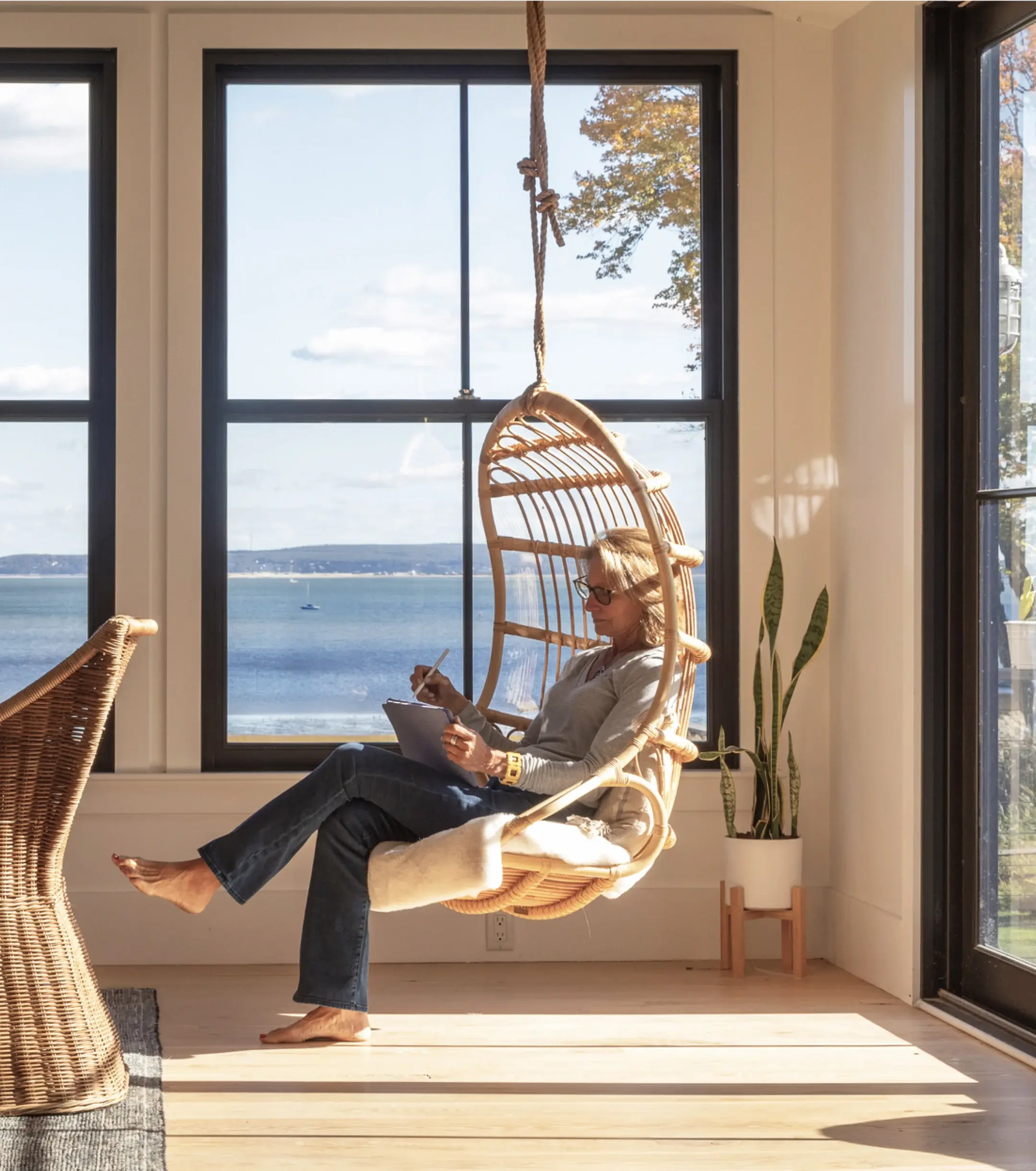 Person sits in a wicker hanging chair by a large window, writing in a notebook. Sunlight fills the room, with a view of the ocean and a flag outside.