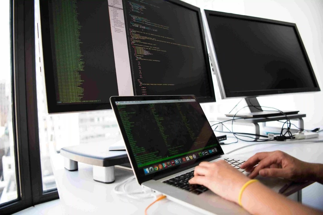 MULTIPLE COMPUTERS ON A DESK WHILE A PERSON IS WORKING ON ONE AND THERE IS A LARGE SKYSCRAPER IN THE BACKGROUND