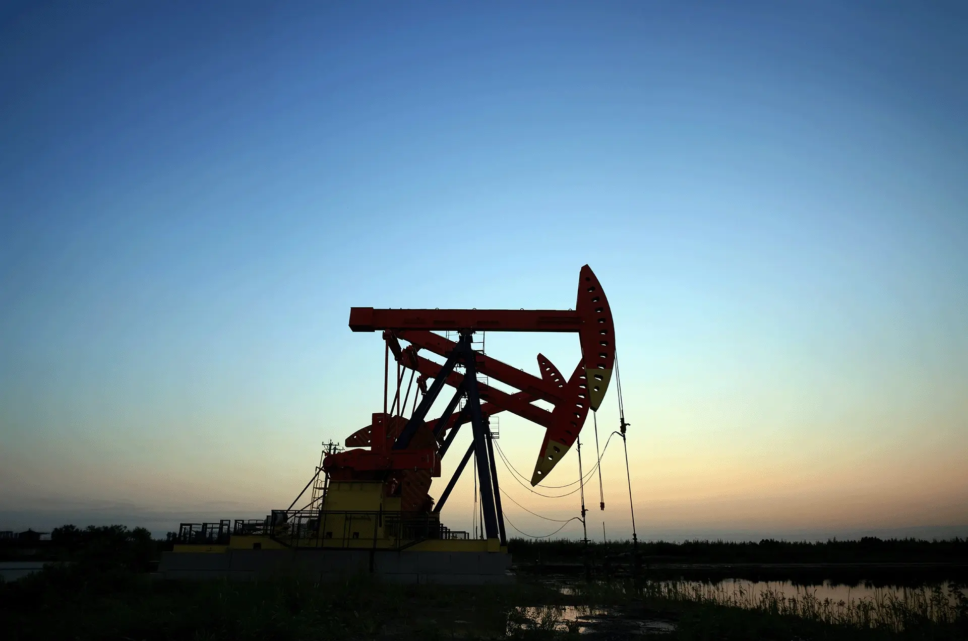 Oil pumpjack silhouetted against a sunset sky beside a reflective body of water.
