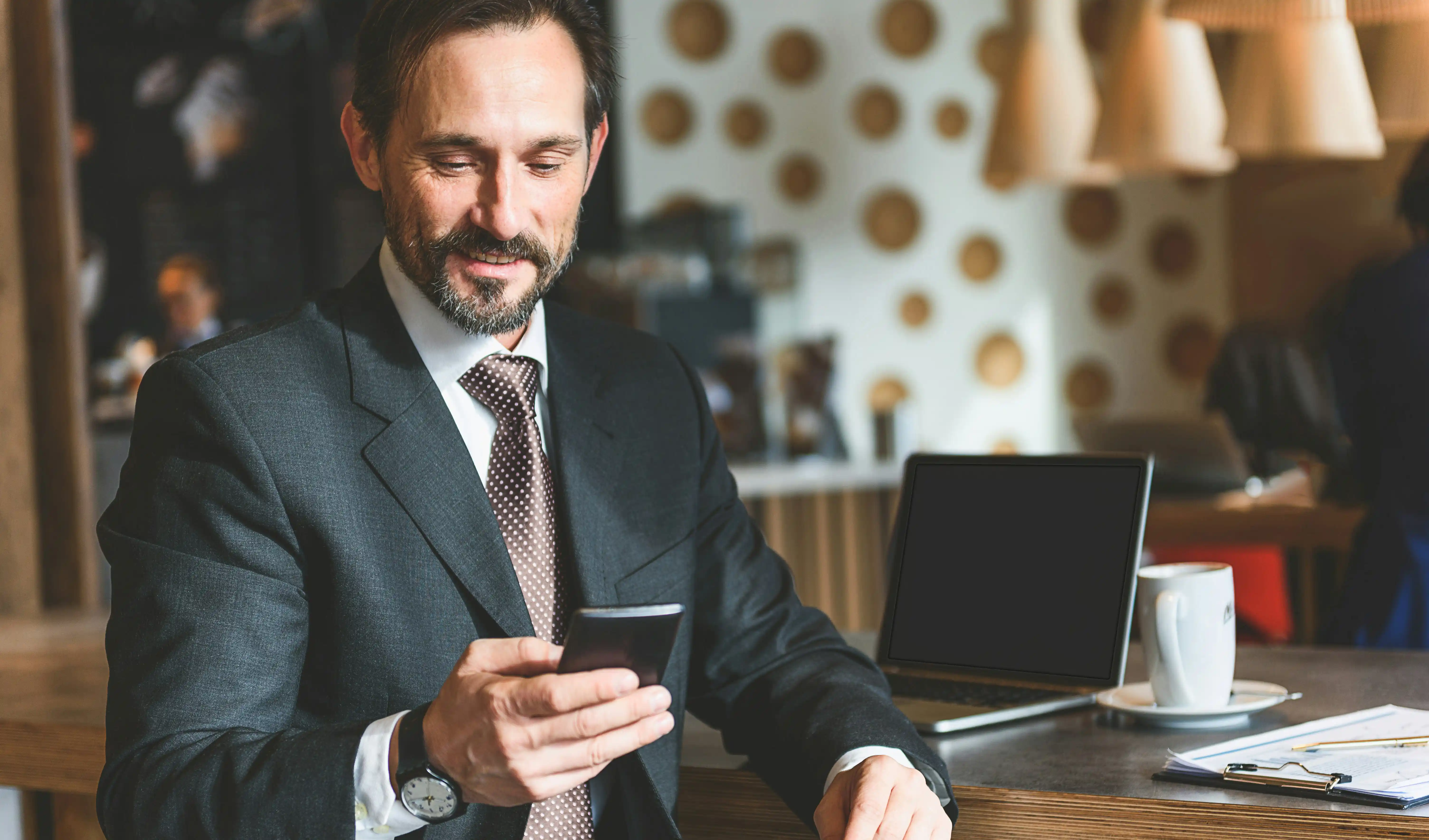 A man in a dark suit and tie sits at a table in a café, smiling while looking at his smartphone, with a laptop and coffee cup beside him.