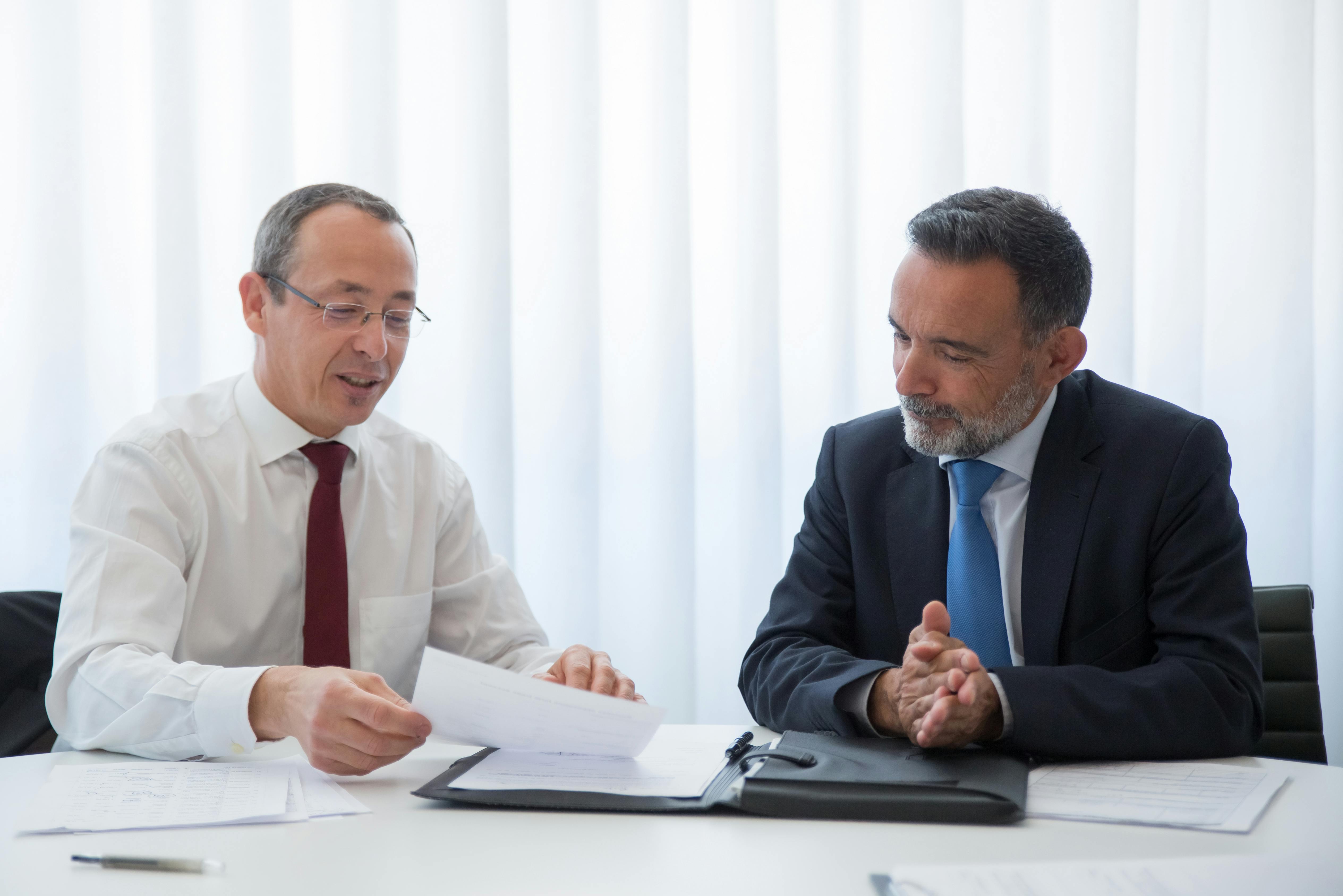 Two men in business attire sit at a table reviewing documents together; one holds a sheet of paper while the other listens attentively with his hands clasped.