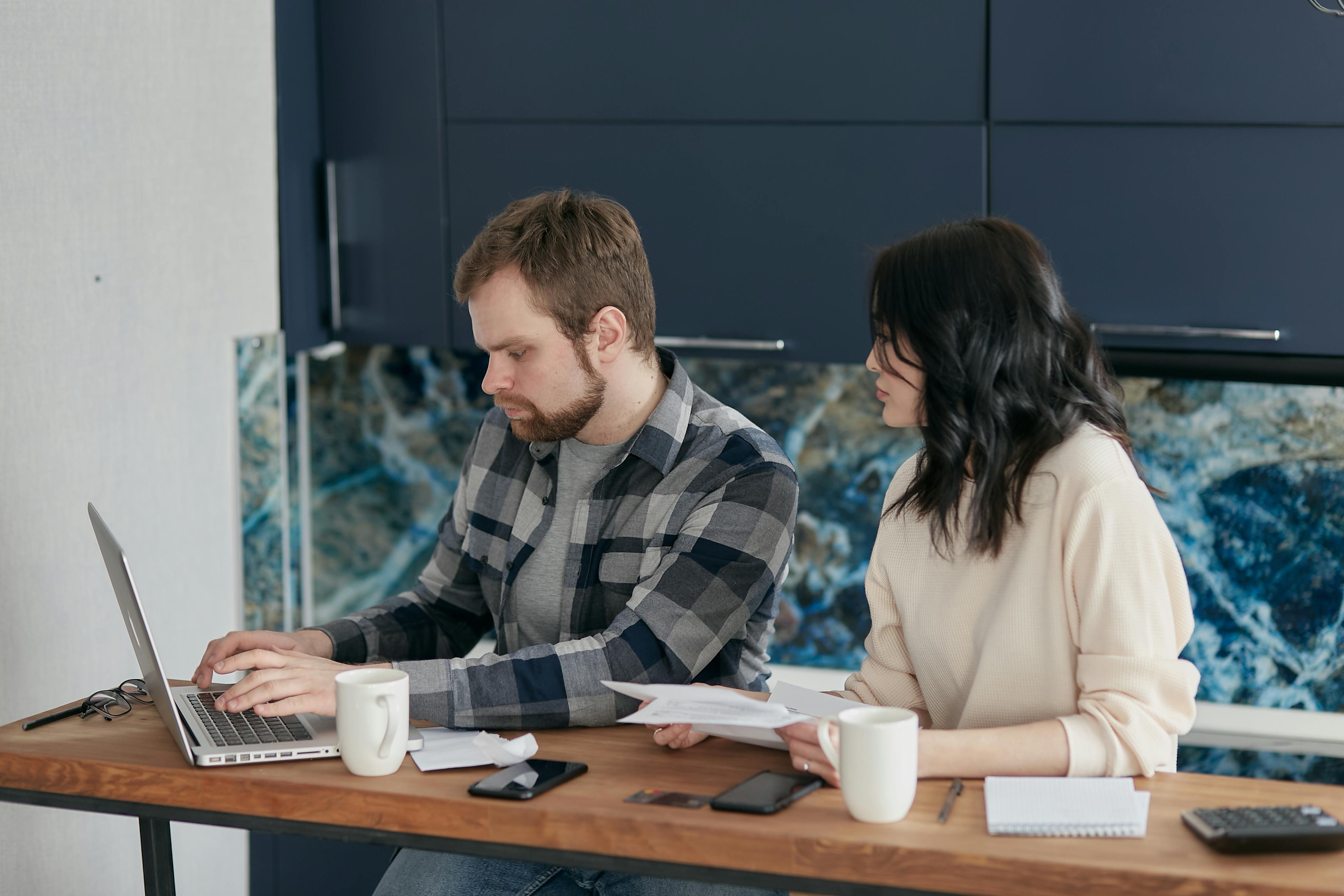 A man and a woman sit at a wooden table covered with papers, phones, notebooks, and coffee mugs. The man is focused on a laptop while the woman, holding documents, looks toward him as if discussing something. They appear to be working together in a modern kitchen with dark cabinets and a blue-patterned backsplash.